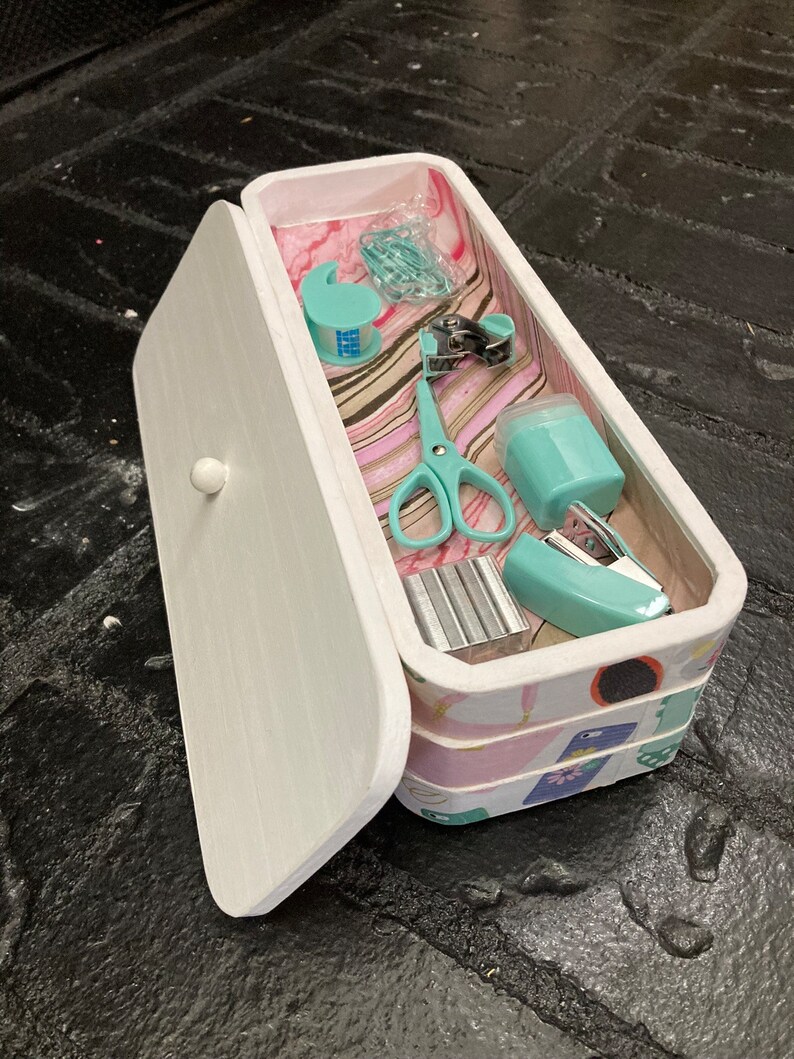 May include: A white, rectangular box with a hinged lid, containing various teal-colored office supplies. The box is decorated with a patterned design and includes scissors, tape dispenser, stapler, and paper clips.