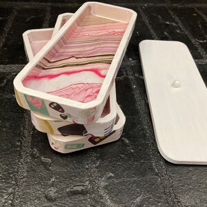 May include: A stack of rectangular trays with a white exterior and a pink, white, and gold marbled interior. The trays have decorative patterns on the sides. A white lid with a small knob is next to the stack.