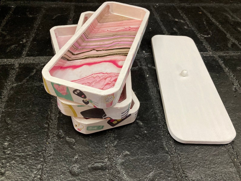 May include: A stack of rectangular trays with a white exterior and a pink, white, and gold marbled interior. The trays have decorative patterns on the sides. A white lid with a small knob is next to the stack.