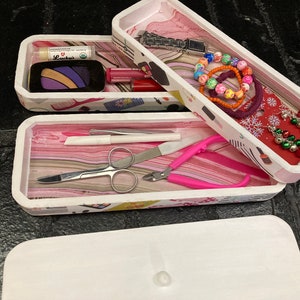 May include: Three white rectangular storage boxes with decorative interiors. The boxes contain various items, including makeup, jewelry, and manicure tools. The interiors feature pink and patterned designs. The lid is white.