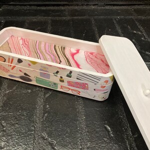 May include: A rectangular wooden box with a white exterior and a patterned interior. The box's exterior is decorated with makeup-related illustrations, while the interior features a pink and white marbled design. The lid is white.