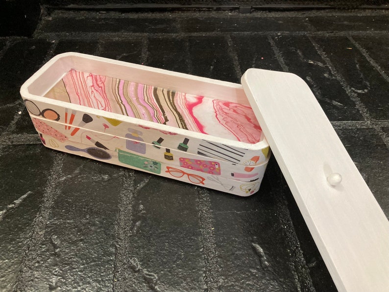 May include: A rectangular wooden box with a white exterior and a patterned interior. The box's exterior is decorated with makeup-related illustrations, while the interior features a pink and white marbled design. The lid is white.