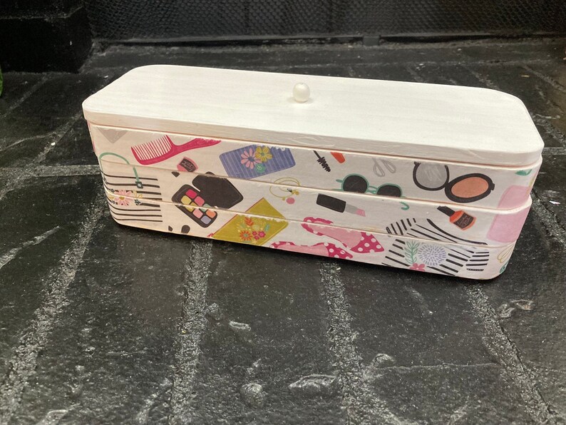 May include: A rectangular white wooden box with a lid and a small knob. The sides are decorated with a repeating pattern of makeup items, including lipsticks, sunglasses, and makeup brushes. The box is designed for storing beauty products.