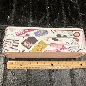 May include: A rectangular wooden box with a light pink base and a patterned top. The top features a repeating design of makeup items, purses, and shoes. The box measures approximately 12 inches in length.