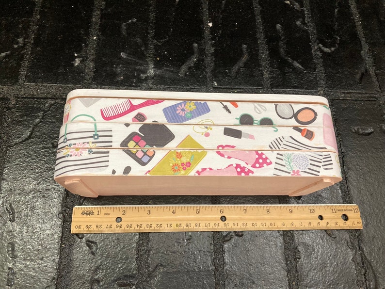 May include: A rectangular wooden box with a light pink base and a patterned top. The top features a repeating design of makeup items, purses, and shoes. The box measures approximately 12 inches in length.