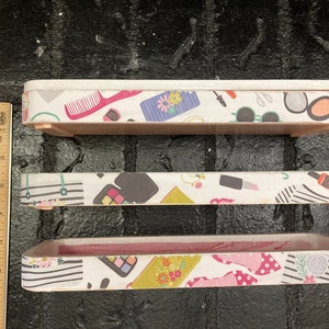 May include: Three rectangular wooden trays with white patterned sides. The patterns include makeup items like combs, sunglasses, and lipstick. The trays are different sizes, with the largest measuring approximately 12 inches long.