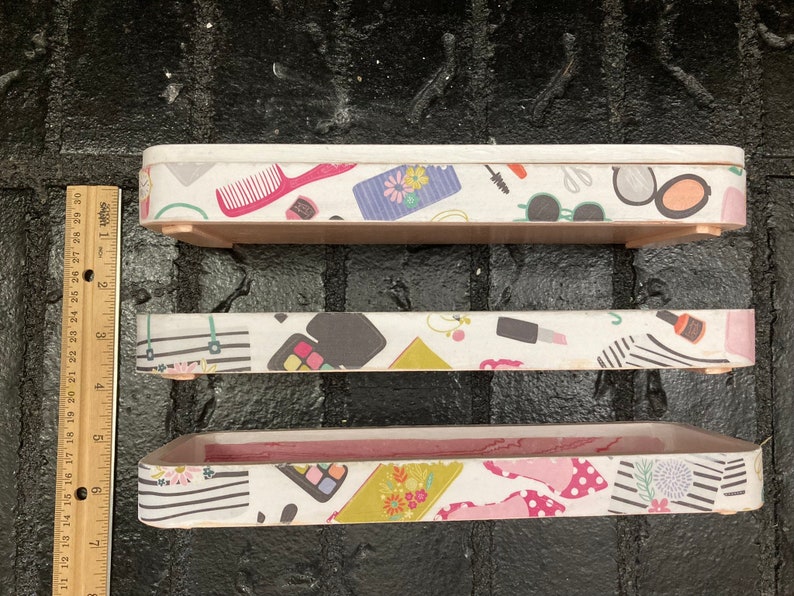 May include: Three rectangular wooden trays with white patterned sides. The patterns include makeup items like combs, sunglasses, and lipstick. The trays are different sizes, with the largest measuring approximately 12 inches long.