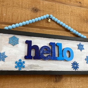 May include: A black framed sign with a white background and the word "hello" in blue letters. The sign is decorated with blue snowflake cutouts.