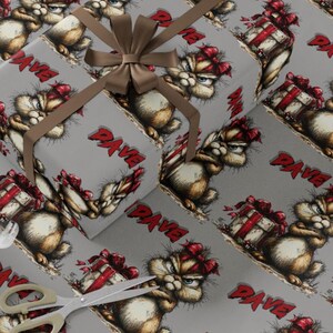 May include: Gray wrapping paper featuring a cartoon cat with a red bow and the name "DAVE" in red. The design includes wrapped gifts with red bows. A brown ribbon and bow are on top of the wrapping paper. Scissors are in the lower left corner.