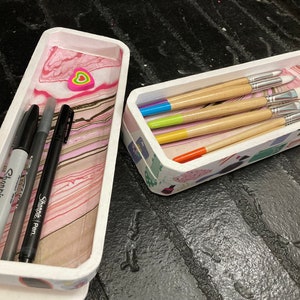 May include: Two rectangular, white trays with decorative edges. One tray contains three black markers and a gray Sharpie marker. The other tray holds paint brushes with colorful handles. The trays sit on a dark, textured surface.