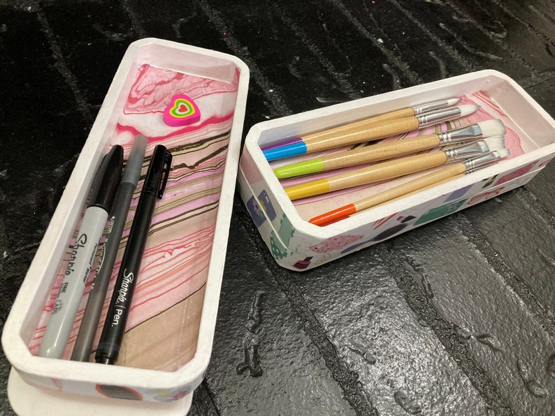 May include: Two rectangular, white trays with decorative edges. One tray contains three black markers and a gray Sharpie marker. The other tray holds paint brushes with colorful handles. The trays sit on a dark, textured surface.