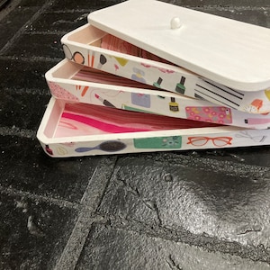 May include: A stackable white wooden storage box set with a lid. The interior of each tray features a different decorative paper design with beauty-related items like makeup, nail polish, and eyeglasses. The trays are stacked on top of each other.