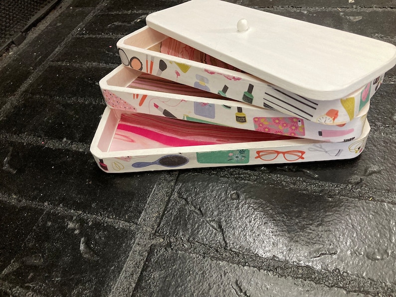 May include: A stackable white wooden storage box set with a lid. The interior of each tray features a different decorative paper design with beauty-related items like makeup, nail polish, and eyeglasses. The trays are stacked on top of each other.