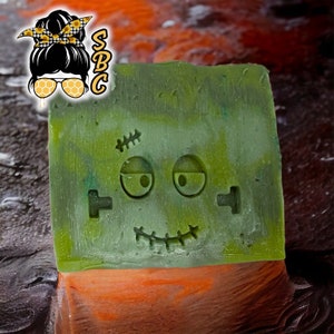 May include: A green, square-shaped soap bar with a carved Frankenstein face. The soap has a stitched mouth, eye sockets, and forehead scars. The soap sits on an orange and black background.