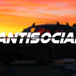 Puede incluir: Un coche negro con una calcomanía blanca "ANTISOCIAL" en la ventana trasera. El coche está aparcado delante de un fondo borroso de puesta de sol.
