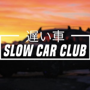 Peut inclure: Silhouette d'une voiture sombre contre un coucher de soleil vibrant. Le texte "SLOW CAR CLUB" est affiché en blanc, avec des caractères japonais au-dessus. L'image a une ambiance cinématographique.