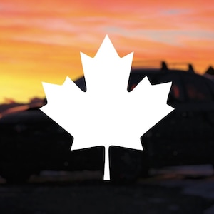 May include: A white maple leaf silhouette is centered in front of a car, set against a vibrant sunset sky. The car is dark, and the background features a blend of orange, yellow, and red hues.