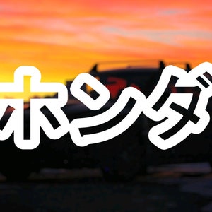 Peut inclure: Un autocollant blanc avec le mot japonais "ホシノ" (Hoshino) sur une voiture noire. La voiture est garée devant un ciel de coucher de soleil.