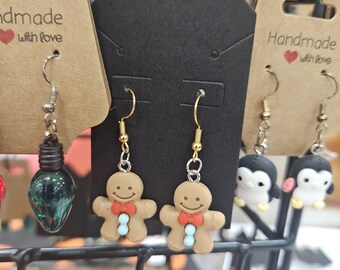 May include: A selection of handmade earrings on display. One pair features a green bulb, another has gingerbread men with red bow ties and blue buttons, and a third pair has penguin charms. The earrings hang from black and tan tags.