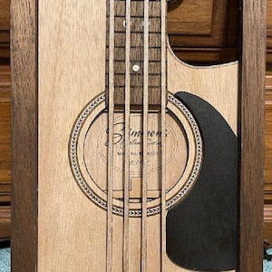 May include: Wooden framed wall art depicting a bass guitar. The artwork features a light brown guitar body with a dark brown fretboard and strings. A black pickguard is present, and the text "Simmons" is visible.