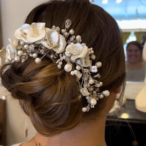 May include: A bridal hair accessory featuring ivory fabric flowers, pearls, and crystal accents. The floral arrangement is attached to a silver wire base and is worn in an updo hairstyle. The accessory adds a touch of elegance.