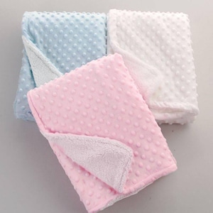 Op de afbeelding: Drie babydekens in pastelkleuren: lichtblauw, roze en wit. Elke deken heeft een gestructureerd, gestippeld oppervlak aan één kant en een zachte, pluche voering aan de andere kant. De dekens zijn opgevouwen en tonen de contrasterende texturen en kleuren.