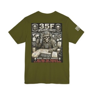May include: An olive green t-shirt featuring a graphic on the back of a skeleton in military gear working at a desk. The text reads 35F INTELLIGENCE ANALYST, SILENCE. ANALYSIS. DOMINANCE. WE FIND THE THREATS SO OTHERS DON'T HAVE TO FIND THE BODIES.