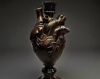 May include: A bronze-colored, heart-shaped sculpture with a dark base and a cylindrical top. The sculpture has detailed veins and anatomical features, and it stands on a pedestal base. The overall design is artistic and unique.