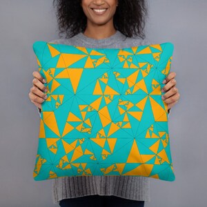 May include: Square throw pillow with a geometric pattern. The pillow features a teal background with a repeating pattern of orange triangles and black lines. The pillow is held up by a person.