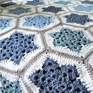 Könnte beinhalten: Eine handgefertigte Häkeldecke mit einem geometrischen Muster. Die Decke hat ein sich wiederholendes Design aus blauen, weißen und grauen sechseckigen Formen mit sternförmigen Zentren. Die Decke ist aus weichem Garn gefertigt und eignet sich für ein Bett oder Sofa.