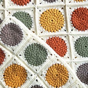 Puede incluir: Una manta de ganchillo hecha a mano con un patrón repetitivo de círculos coloridos. Los círculos son en tonos de naranja, amarillo, verde y gris, sobre un fondo crema. La manta está hecha de hilo suave y tiene una apariencia texturizada.