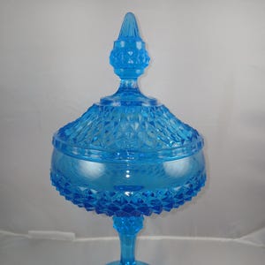 May include: A vintage, light blue glass candy dish with a lid. The dish has a diamond pattern on the lid and bowl, and sits on a pedestal base. The finial on the lid is pointed. The candy dish is a decorative piece.