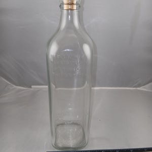 May include: A clear glass bottle with a rectangular body and a cork and red cap. The bottle has embossed text and a rounded shoulder. A ruler is at the bottom of the image.