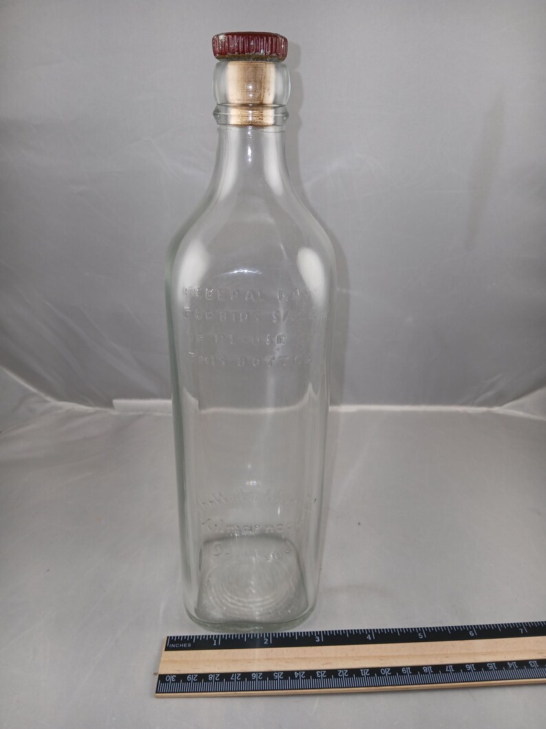 May include: A clear glass bottle with a rectangular body and a cork and red cap. The bottle has embossed text and a rounded shoulder. A ruler is at the bottom of the image.