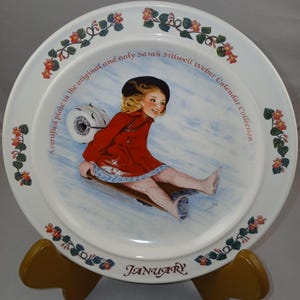 May include: A white ceramic plate with a floral border and a picture of a girl in a red coat sledding down a snowy hill. The plate says "A certified place in the original and only Sarah Stilwell Weber Calendar Collection" and "JANUARY".