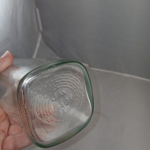 May include: Clear glass bottle with a square base and rounded corners. The bottom of the bottle has a circular design with embossed text. The bottle is held by a hand.