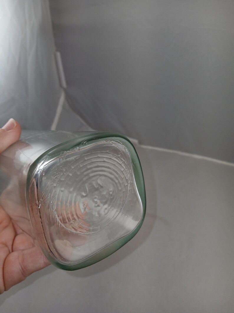 May include: Clear glass bottle with a square base and rounded corners. The bottom of the bottle has a circular design with embossed text. The bottle is held by a hand.