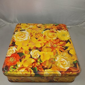 May include: A rectangular metal tin featuring a bright floral pattern. The lid and sides are adorned with yellow, orange, and white flowers, including roses. The tin is empty and appears to be made of metal.