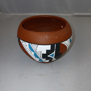 Puede incluir: Un cuenco redondo de cerámica color terracota con una abertura ancha. El cuenco presenta un diseño geométrico en negro, blanco y azul alrededor del exterior. El interior del cuenco es de un tono marrón más oscuro.