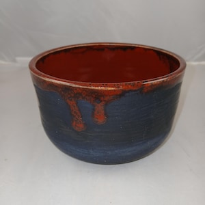 Puede incluir: Un cuenco de cerámica hecho a mano con exterior azul oscuro e interior marrón rojizo. El borde tiene un efecto de esmalte texturizado y goteado. La forma del cuenco es redonda y ligeramente cónica, adecuada para servir o exhibir.