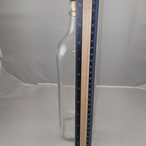 May include: A clear glass bottle with a red and gold stopper. A ruler is placed next to the bottle, showing a height of approximately 10 inches. The bottle has a rectangular shape and a narrow neck.