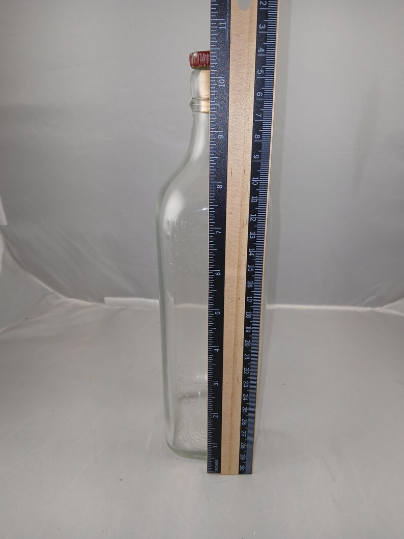 May include: A clear glass bottle with a red and gold stopper. A ruler is placed next to the bottle, showing a height of approximately 10 inches. The bottle has a rectangular shape and a narrow neck.