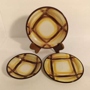 Puede incluir: Tres platos de cerámica con un diseño geométrico marrón y amarillo. El plato más grande está en un soporte de madera. Los platos más pequeños están en una superficie blanca.