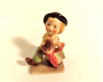 May include: A small porcelain figurine of a child with rosy cheeks, wearing a black hat, green pants, and a red scarf, holding a brown dog. The figurine is on a rectangular base.