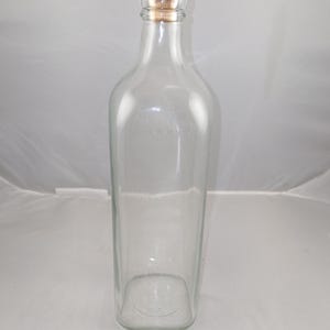 May include: A clear glass bottle with a rectangular base and a cork stopper. The stopper has a red top and a natural cork base. The bottle is empty and has a simple, elegant design.