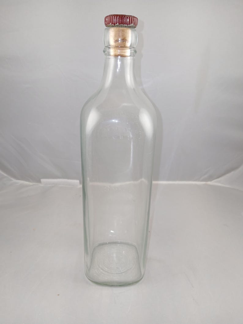 May include: A clear glass bottle with a rectangular base and a cork stopper. The stopper has a red top and a natural cork base. The bottle is empty and has a simple, elegant design.