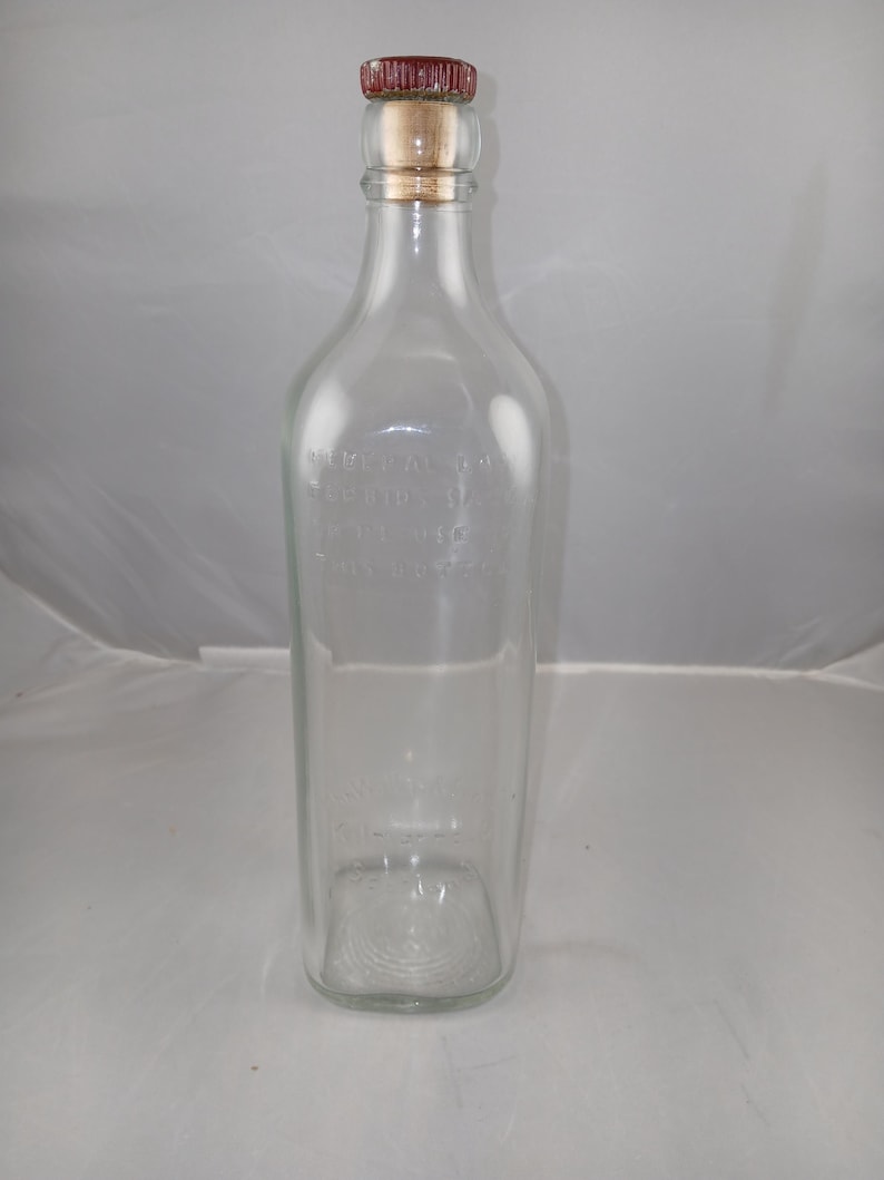 May include: A clear glass bottle with a red and tan cap. The bottle has a rectangular base and a long, slender neck. The bottle has text on the front. The cap is made of a cork-like material and is topped with a red, textured material.
