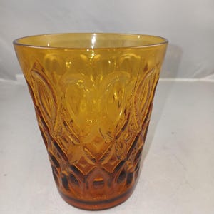 May include: A translucent amber-colored glass tumbler featuring a raised, repeating geometric pattern across its exterior surface. The vessel has a wide, circular rim that tapers down to a narrower base, showcasing a textured, pressed glass design.