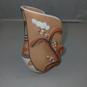May include: A handcrafted ceramic vase with a unique, asymmetrical shape. The vase features a tan base color with black, orange, and white painted designs. Decorative elements include cloud-like shapes and abstract floral patterns. The vase has a curved opening.
