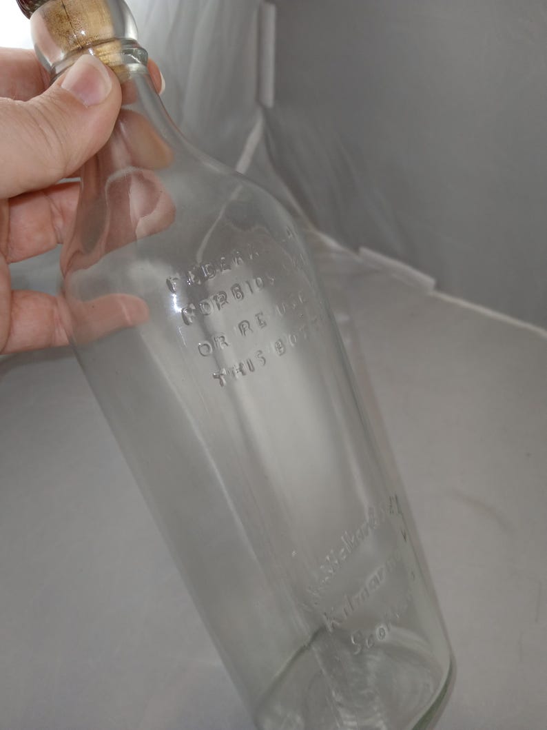 May include: A clear glass bottle with a wooden stopper. The bottle has embossed text that reads "ILLEGAL FORBIDDEN OR RE USE THIS BOTTLE" and "J. Walker & Co. Kilmarnock Scotland".